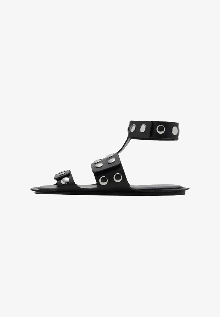 Sandale negre plate cu multiple bretele și cuie circulare din argint. Prezintă un design elegant și o textură netedă. Are spatele și zona de degetele deschise.