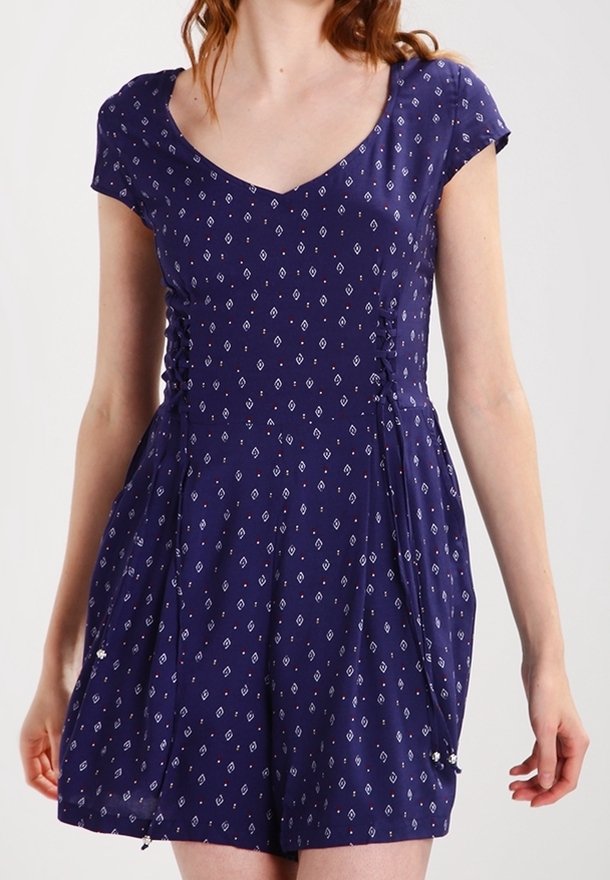 Femme portant une robe bleu marine à manches courtes avec un petit motif losange blanc et des côtés froncés avec des panneaux en tissu plissé.