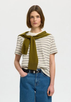 Ung person med kort brunt hår iført en hvid og sort stribet T-shirt, blå jeans, sort bælte og en olivengrøn sweater draperet over skuldrene.