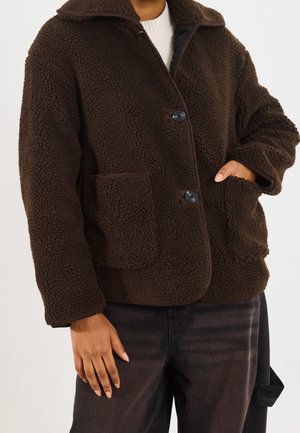 Personne portant une veste boutonnée marron foncé duveteuse avec des poches avant, associée à un pantalon foncé, debout devant un fond uni.