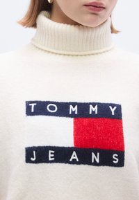 Pull col roulé crème avec texture côtelée, arborant un patch logo rectangulaire bleu marine et rouge où est inscrit "TOMMY JEANS" en lettres blanches.