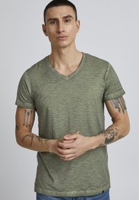 Maslinasto zelena majica s v-neck dekolteom od teksturirane, lagane tkanine. Kratki rukavi i minimalni detalji šivanja. Model s tetovažama pozira s rukama sklopljenim.