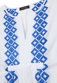 Blusa in tessuto bianco con ricami geometrici blu e motivi a diamante sul davanti e scollatura profonda a V. L'etichetta riporta "PIOMBO."
