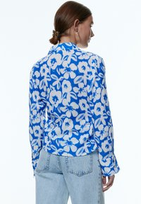 Blumenbluse in Blau und Weiß, aus leichtem Stoff mit langen Ärmeln und einem kragenähnlichen Halsbereich, abgerundetem Saum und geknöpften Bündchen.