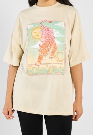 Beżowy T-shirt z kolorową grafiką tygrysa, słońca, księżyca, gwiazd oraz tekstem "Stay Wild", noszony przez osobę w czarnych spodniach.