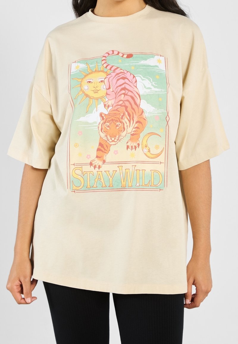 T-shirt beige avec un graphisme coloré de tigre, soleil, lune, étoiles et texte "Stay Wild" porté par une personne portant un pantalon noir.