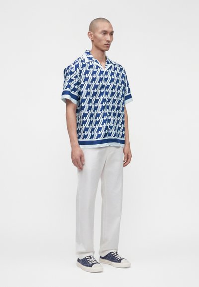 Hombre con camisa de manga corta azul y blanca con estampado, pantalones blancos y deportivas azul marino, de pie frente a un fondo blanco liso.