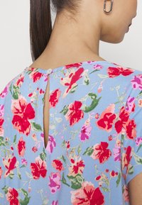 Donna con un lungo ponytail scuro che indossa un vestito floreale blu con fiori rossi, rosa e verdi, caratterizzato da uno spacco sul retro fissato da un piccolo bottone.