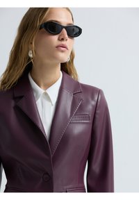 Blazer in pelle bordeaux con rever a punta, dettagli di cucitura sottili e chiusura a un bottone, indossato sopra una camicia bianca.
