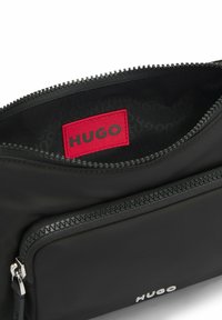 Borsetta nera con zip e superficie opaca; presenta un'etichetta rossa "HUGO" e un piccolo scomparto con zip sulla parte anteriore.