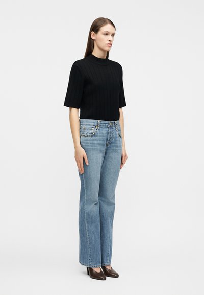 Γυναίκα που στέκεται φορώντας μαύρο ριμπ τοπ με κοντά μανίκια, ανοιχτό μπλε τζιν με ίσια μπατζάκια και σκοτεινές μυτερές γόβες μπροστά σε απλό λευκό φόντο.