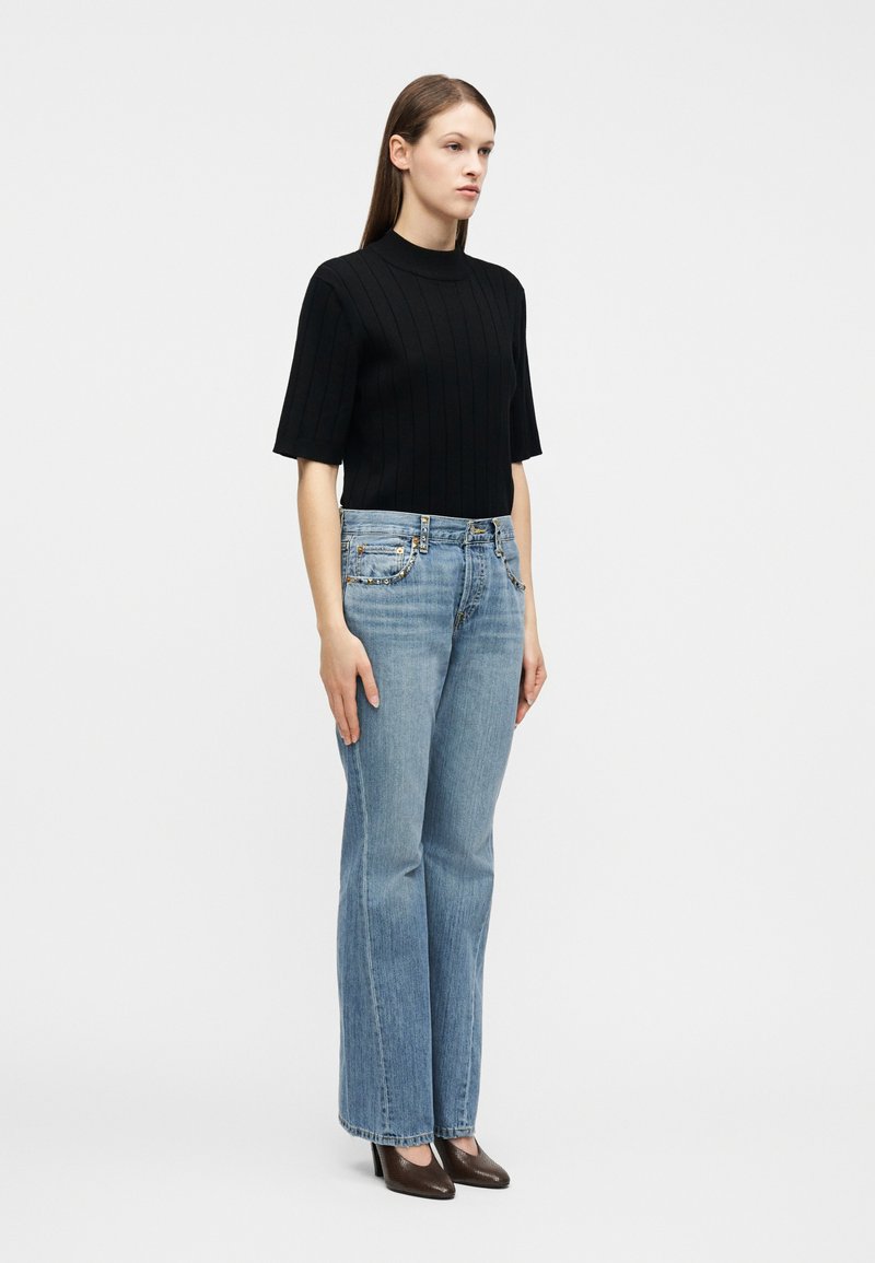 Kvinde stående iført en sort kortærmet ribbet top, lyseblå jeans med lige ben og mørke spidse hæle mod en ensartet hvid baggrund.