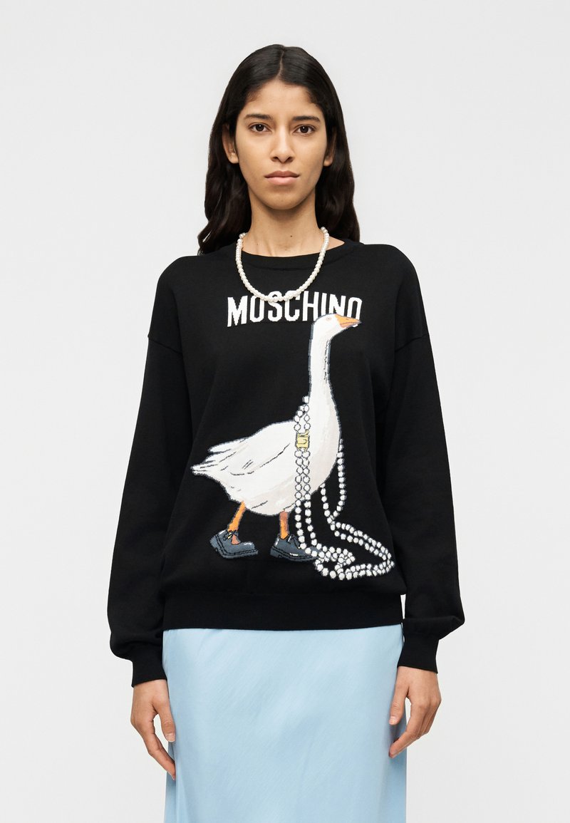 Μαύρο πουλόβερ με λευκό σχέδιο χήν ας που φοράει κολιέ από πέρλες και μαύρα παπούτσια. Πάνω από το σχέδιο είναι τυπωμένη η λέξη "MOSCHINO". Μαλακή υφή.