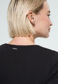 Femme blonde aux cheveux courts portant un haut noir en côtes, laissant découvrir le cou et avec plusieurs piercings en argent aux oreilles, sur un fond clair.