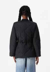Veste matelassée noire avec un motif ondulé, présentant une taille cintrée et un col côtelé. Les manches longues et une coupe décontractée complètent le design.