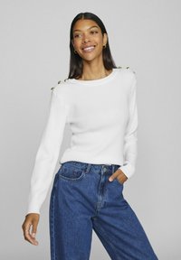 Witte geribde trui met lange mouwen en gouden knoopaccenten op de schouders, gecombineerd met blauwe hooggetailleerde jeans. Soepele textuur en casual pasvorm.