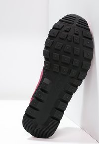 Svart gummisul av Nike-skor med fyrkantiga mönster och övre del i rosa tyg mot en vit hörnbakgrund.