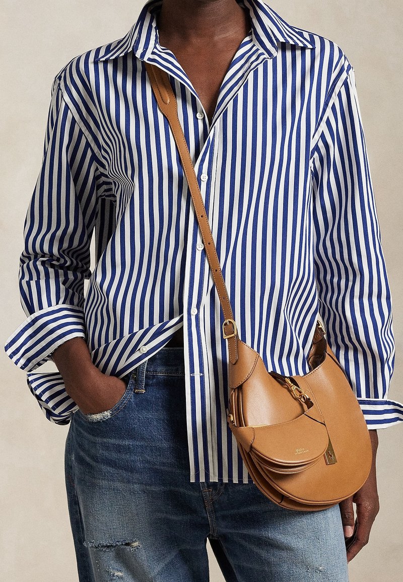 Personne portant une chemise rayée verticale bleue et blanche, un jean bleu, et portant un sac à bandoulière en cuir marron avec une boucle.