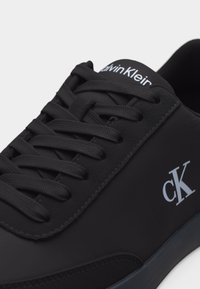 Gros plan sur une basket noire Calvin Klein montrant les lacets, la languette avec la marque et le logo "cK" sur le côté.