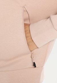 Felpa rosa chiaro con tasca con zip e polsini a costine, realizzata in tessuto morbido con una texture liscia, che mette in evidenza il design funzionale.