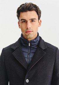 Czarny i granatowy płaszcz w kratę z wysokim kołnierzem i zapinaną na zamek podszewką. Posiada duże guziki i teksturowany materiał. Klasyczny design.