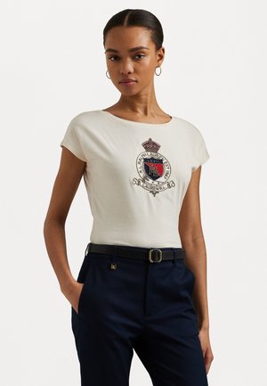 T-shirt crema a maniche corte con un design di stemma ricamato nella parte anteriore, abbinato a pantaloni blu navy e una cintura nera.
