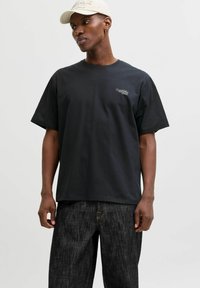 Schwarzes Baumwoll-T-Shirt mit kurzen Ärmeln und rundem Ausschnitt. Mit weißem, auf der linken Brustseite gesticktem Logo. Kombiniert mit dunklen Jeans.