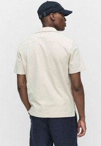 Beżowa koszula z krótkim rękawem, z kołnierzykiem i bocznymi rozcięciami, zestawiona z granatowymi spodniami. Czarna czapka dopełnia całość, widok z tyłu.