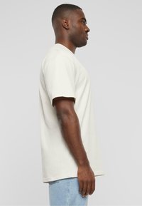 Beige bomull T-skjorte med kort erme og rund hals, vist på en modell fra siden. Løs passform og jevn tekstur.