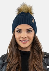 Marineblaue Strickmütze mit Rippstruktur, die oben einen flauschigen Kunstfell-Pompom und seitlich ein kleines markiertes Lederpatch hat.