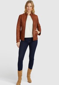 Braune Lederjacke mit hohem Kragen, Reißverschluss und Seitentaschen, kombiniert mit einem weißen Oberteil und dunklen Jeans. Weiche Textur und taillierte Passform.