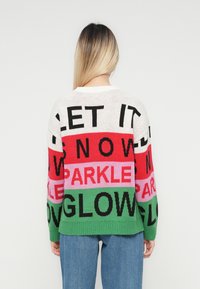 Person mit langen blonden Haaren, die blaue Jeans und einen mehrfarbigen Strickpullover mit dem Text "LASS ES SCHNEIEN, FUNKELN, GLÄNZEN" auf dem Rücken trägt.