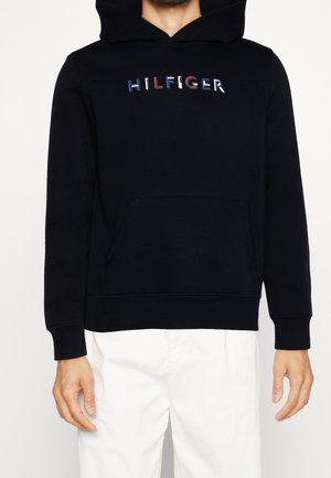 Sudadera negra con bolsillo frontal, que presenta el logo "HILFIGER" multicolor. Fabricada en tejido suave con puños y dobladillo de canalé.