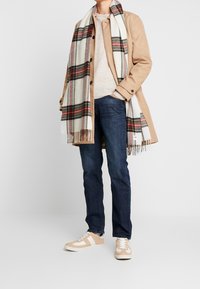 Abrigo beige sobre un suéter beige, jeans oscuros y zapatillas claras con acentos blancos; una bufanda de cuadros multicolores cuelga alrededor del cuello.