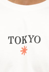 Λευκό T-shirt με μαύρο κείμενο "TOKYO" και ένα κόκκινο αστερίσκο τοποθετημένο στο κέντρο κάτω από το κείμενο στην περιοχή του στήθους.