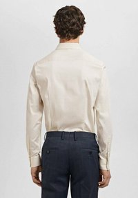 Cămașă lungă bej deschis, cu guler ascuțit, textură netedă, cu manșete cu buton, asociată cu pantaloni închis colorați cu un model, care au două buzunare spate.