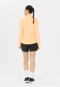 Helles orange Langarmoberteil mit Daumenlöchern, kombiniert mit schwarzen Shorts. Das Model zeigt ein geschichtetes Design, atmungsaktiven Stoff und weiße Socken.