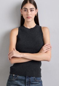 Ung kvinna med långt mörkt hår, bärande guldörhängen, en ärmlös mörkgrå topp och blå jeans, stående med armarna i kors.