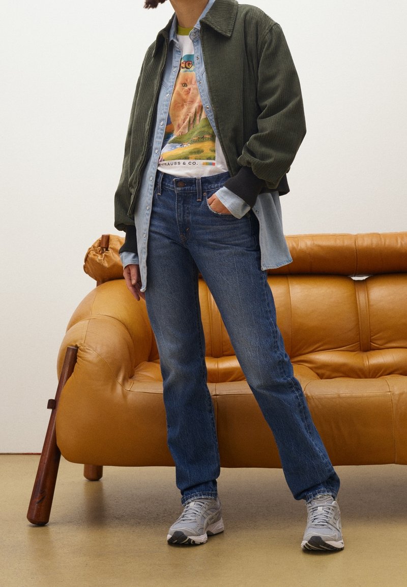 Personne portant un t-shirt à motif sous une chemise en jean et une veste vert olive, un jean bleu et des baskets grises, debout près d'un canapé en cuir beige.