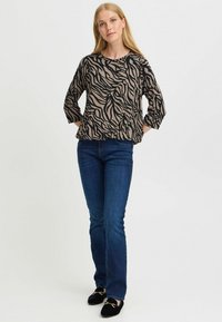 Beige långärmad topp med svart zigzagmönster, avslappnad passform, bärs med mörkblå utsvängda jeans och svarta ballerinas med guld accenter.