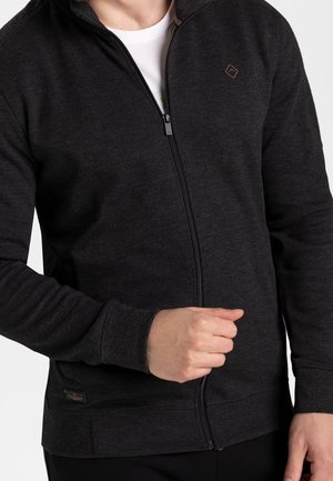 Mężczyzna w ciemnoszarej bluzie rozpinanej na zamek, na białej koszulce, z zamkiem częściowo zamkniętym i prawą ręką blisko zamka.