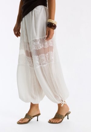Femme portant un pantalon blanc en dentelle transparente avec des bas élastiques et des sandales à talons vert olive, montrée de la taille vers le bas sur fond blanc.