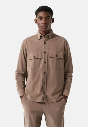 Ung mand iført en lysebrun skjorte med knapper og to brystlommer samt matchende bukser, stående med hænderne i lommerne.