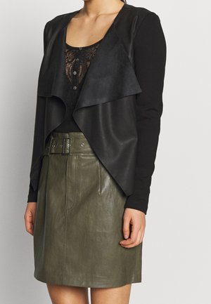 Femme portant un haut noir à manches longues avec un détail en dentelle sur le devant, un gilet noir drapé à ouverture avant et une jupe taille haute ceinturée de couleur vert olive.