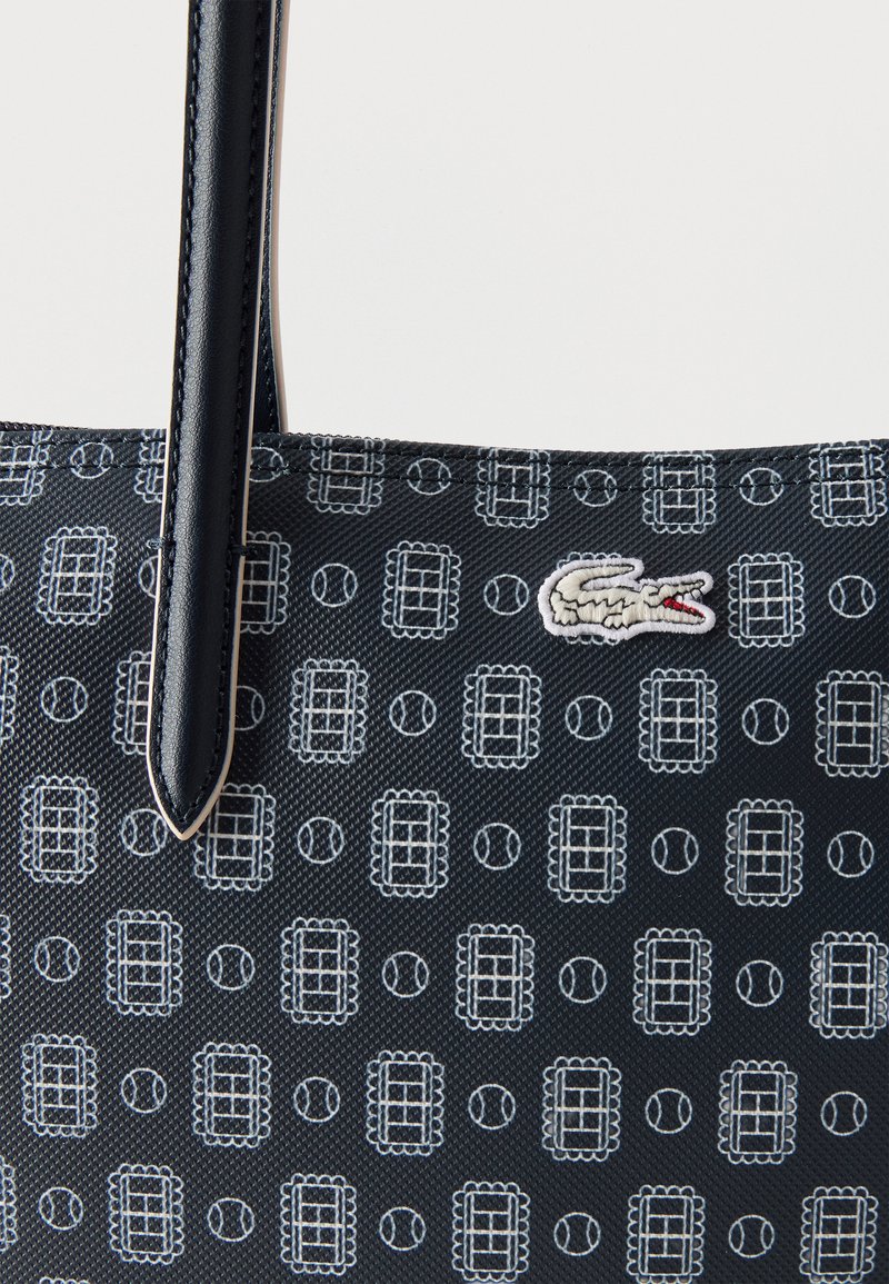 Nahaufnahme einer marineblauen Tragetasche mit einem Muster aus Tennisplatz und Tennisball, bestickt mit Krokodil-Logo und schwarzem Ledergriff.