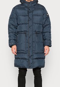 Marinblå dunjacka med huva, som har quiltade sektioner, dragsko i midjan och tryckknappar. Materialet verkar vattentåligt med en slät yta.