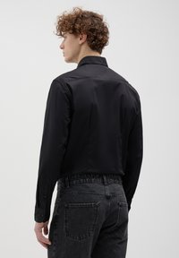 Maglietta nera a maniche lunghe con colletto dritto e polsini con bottone, realizzata in un materiale liscio. Indossata con jeans grigio scuro e texture.