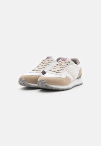 Scarpe da ginnastica beige e bianche realizzate in materiali scamosciati e in rete, con una suola grigia testurizzata e lacci, e un piccolo dettaglio della bandiera di un paese.