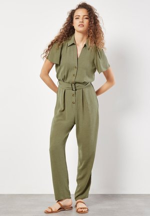 Olivgrüner Jumpsuit mit kurzen Ärmeln, Knopfverschluss, tailliertem Bund und strukturiertem Stoff. Getragen mit beigen flachen Sandalen.
