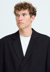 Junger Mann mit lockigem Haar, der einen schwarzen Blazer über einem weißen Hemd trägt und leicht zur Seite schaut, vor einem schlichten hellen Hintergrund.